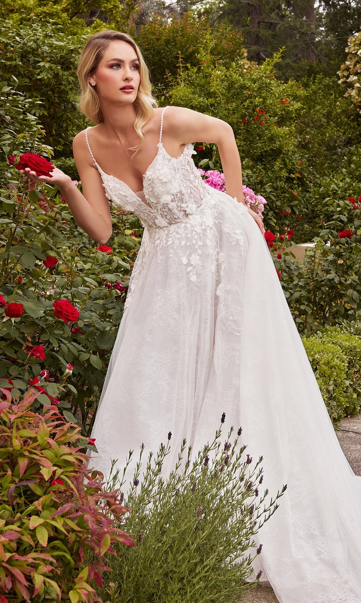 A woman wearing the A-Line White Lace Wedding Gown CD857W with 3D floral lace details stands in a garden, holding a red rose amidst lush greenery and blooming flowers.